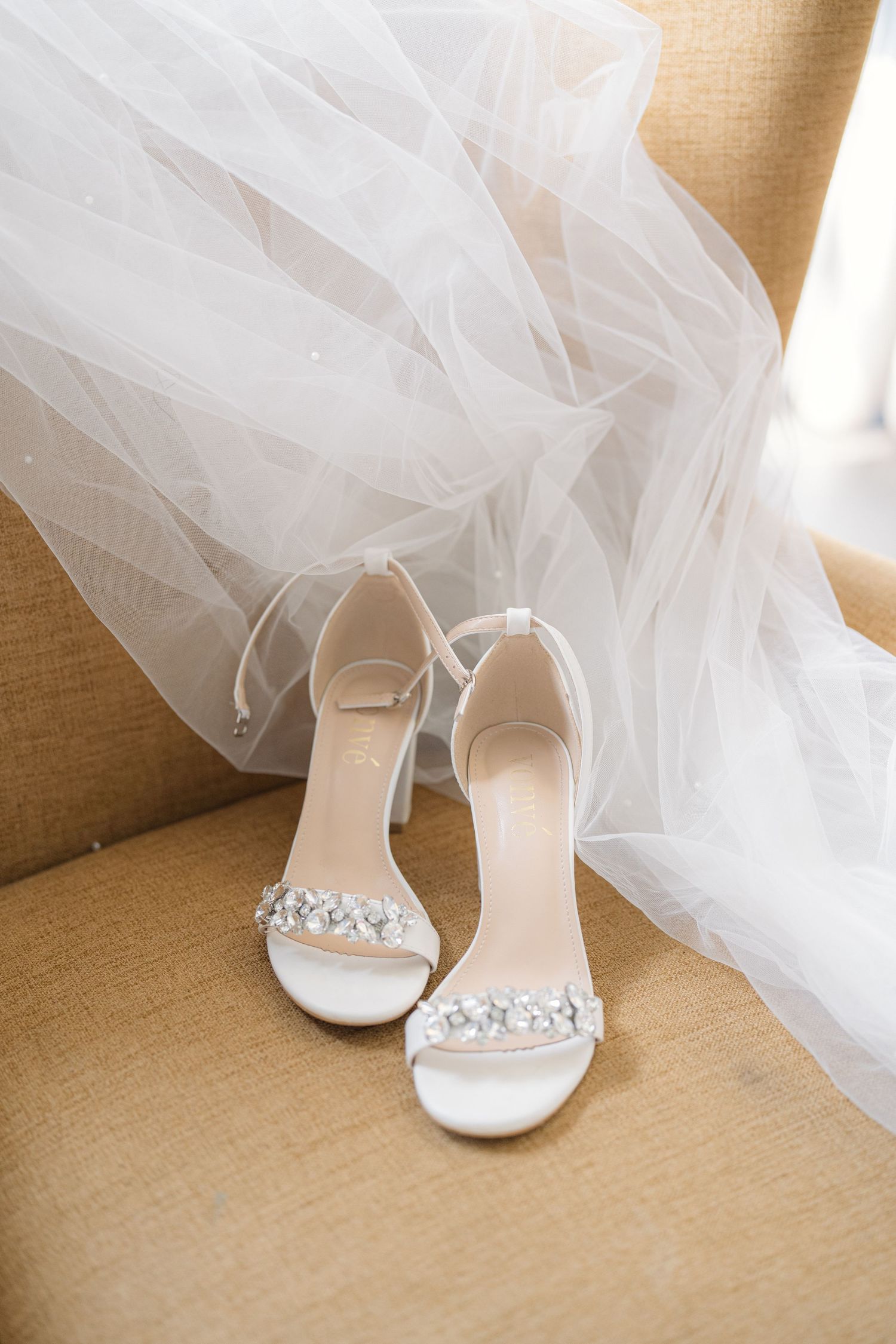 Elegant white wedding shoes adorned with crystal details peek out from beneath a flowing tulle veil on beige carpet.