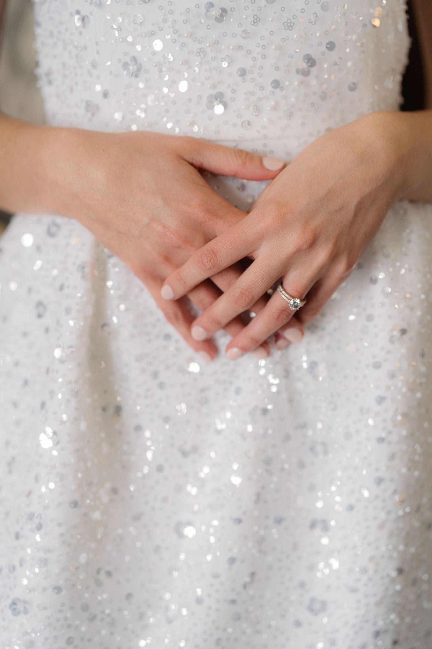 A sparkling white sequined dress fabric with hands showing an engagement ring against the glittery background.