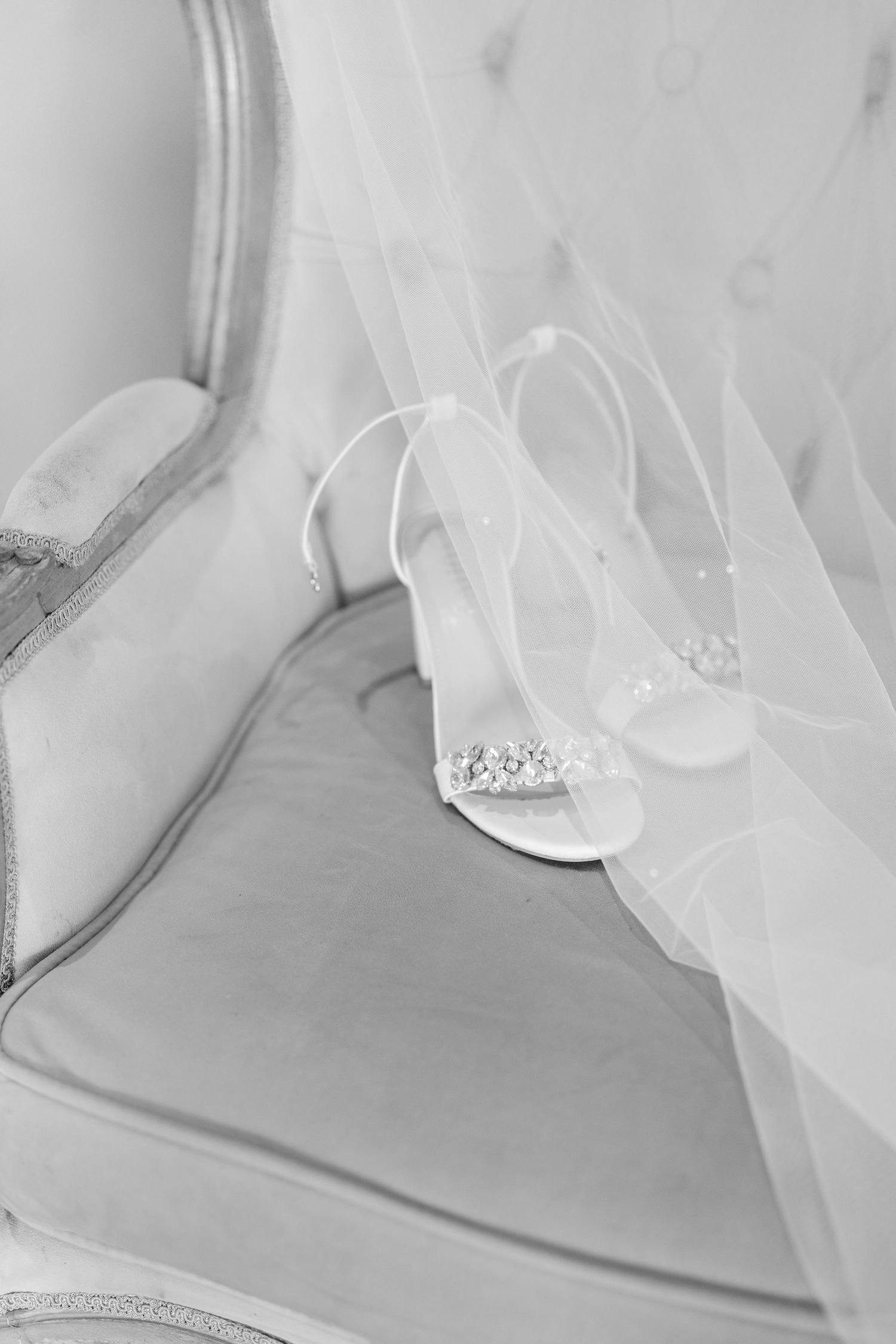 Elegant white bridal shoes with crystal embellishments rest beside flowing tulle veil in black and white photography.