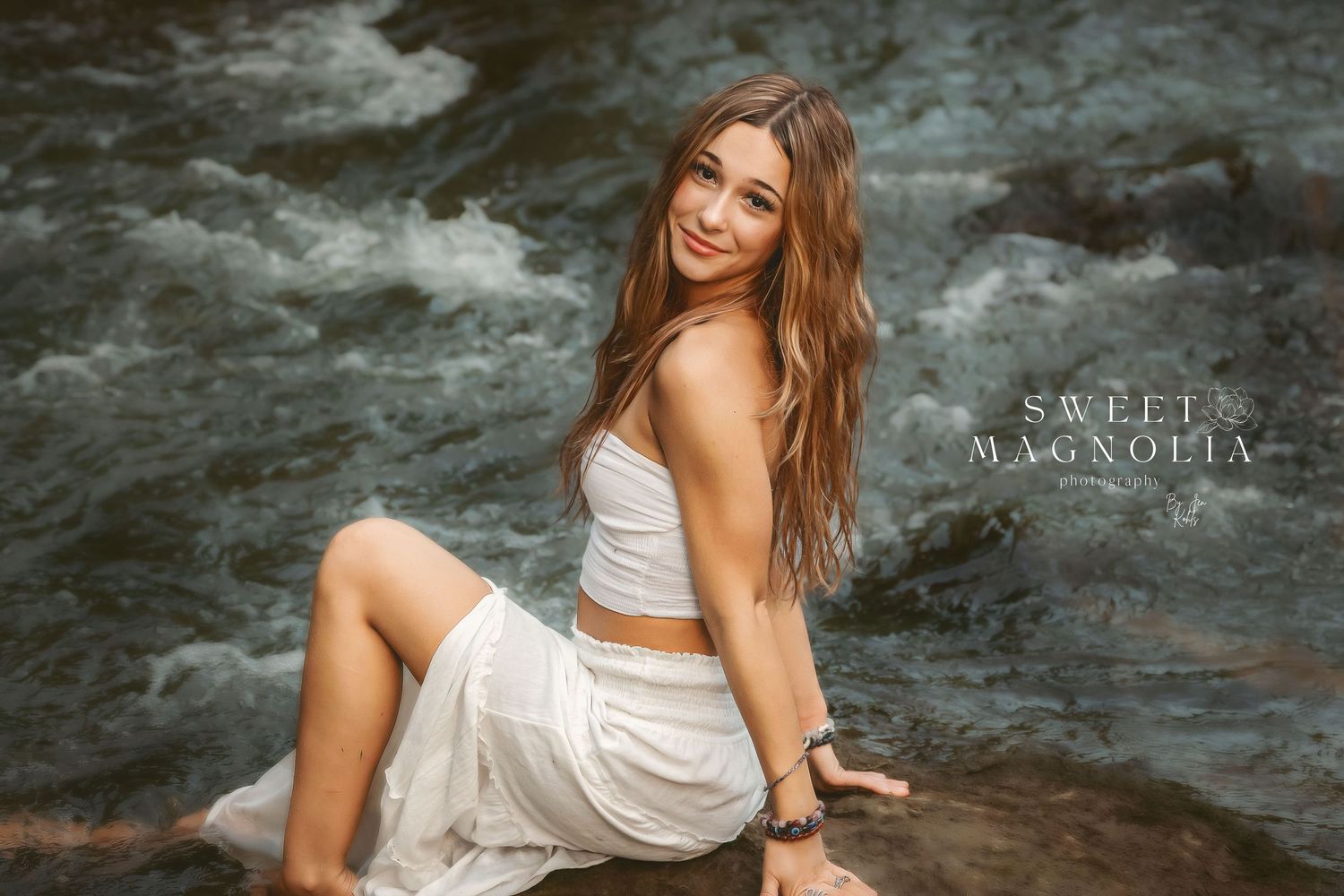 Model in flowing white dress poses by rushing stream in nature setting.