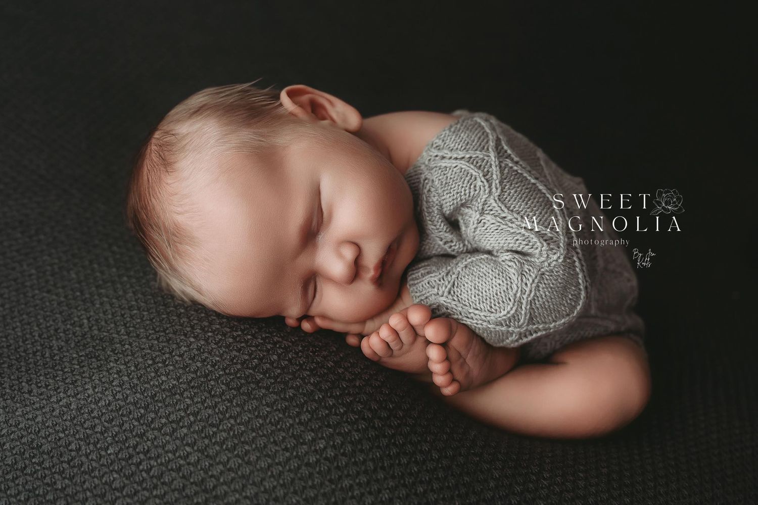 A sleeping newborn baby wrapped in a soft gray blanket lies peacefully on a dark textured surface.