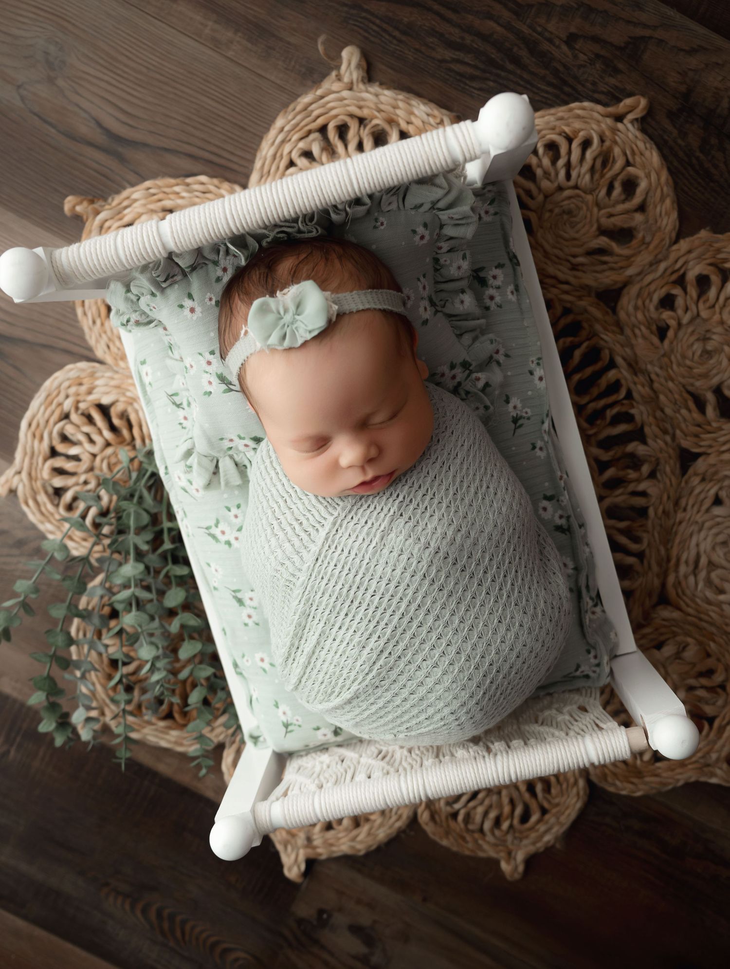 Newborn sleeping peacefully in white swaddle on mint green hammock with ornate woven backdrop.