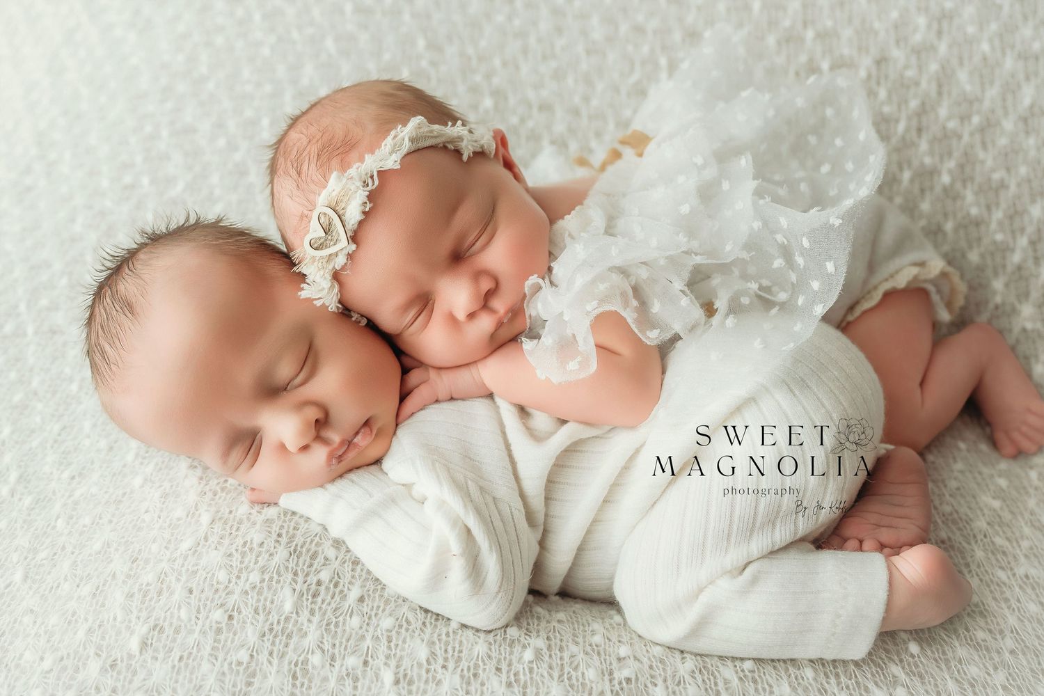 Two sleeping newborns in white outfits lying close together on textured white fabric.