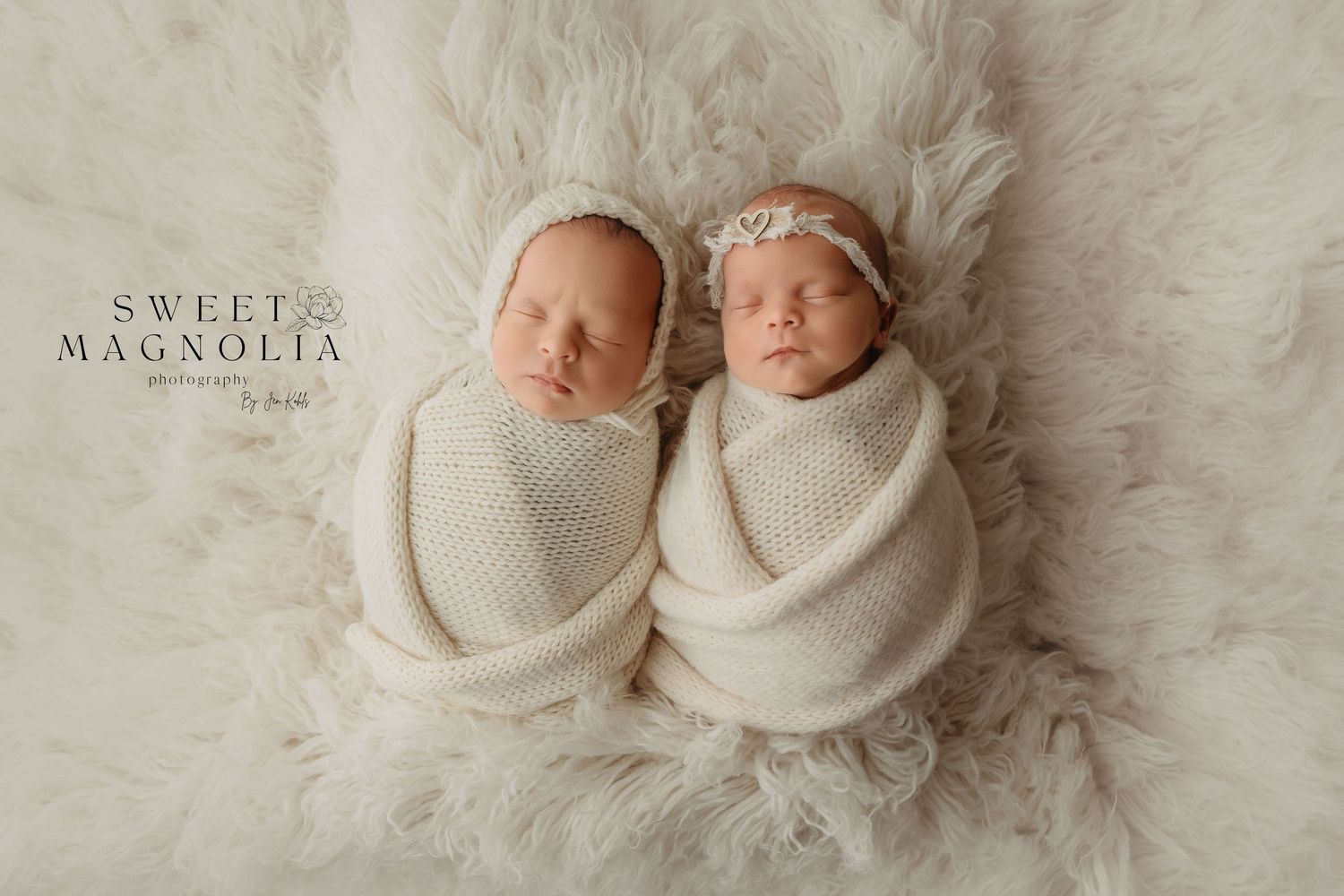 Two sleeping newborns swaddled in cream wraps on soft white fur backdrop.