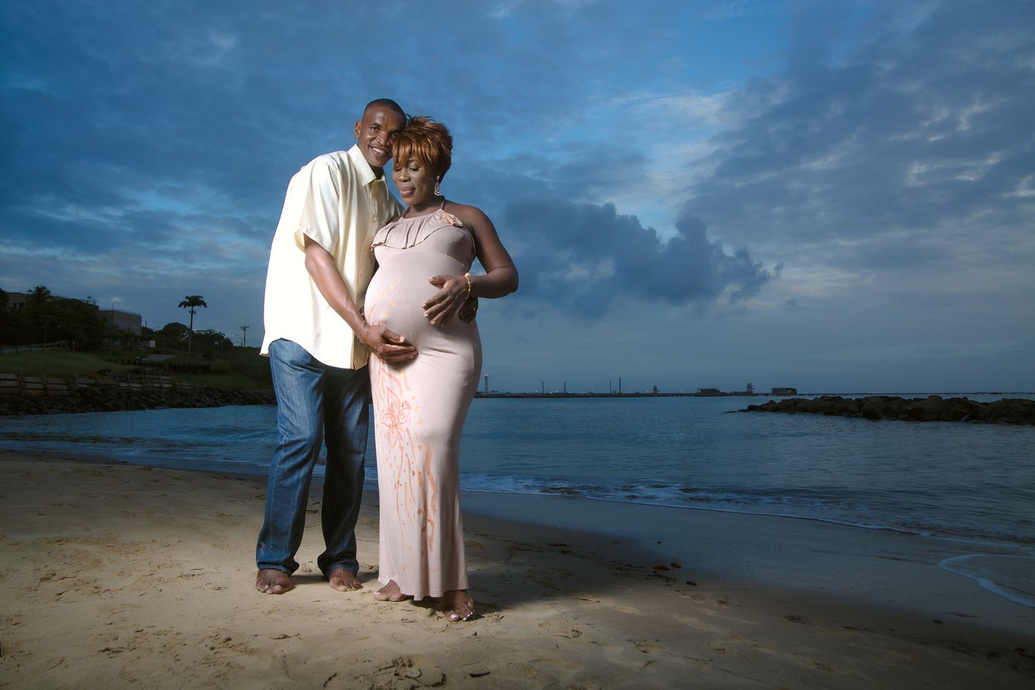 A couple shares an intimate moment on a beach at dusk with dramatic cloudy skies in the background.