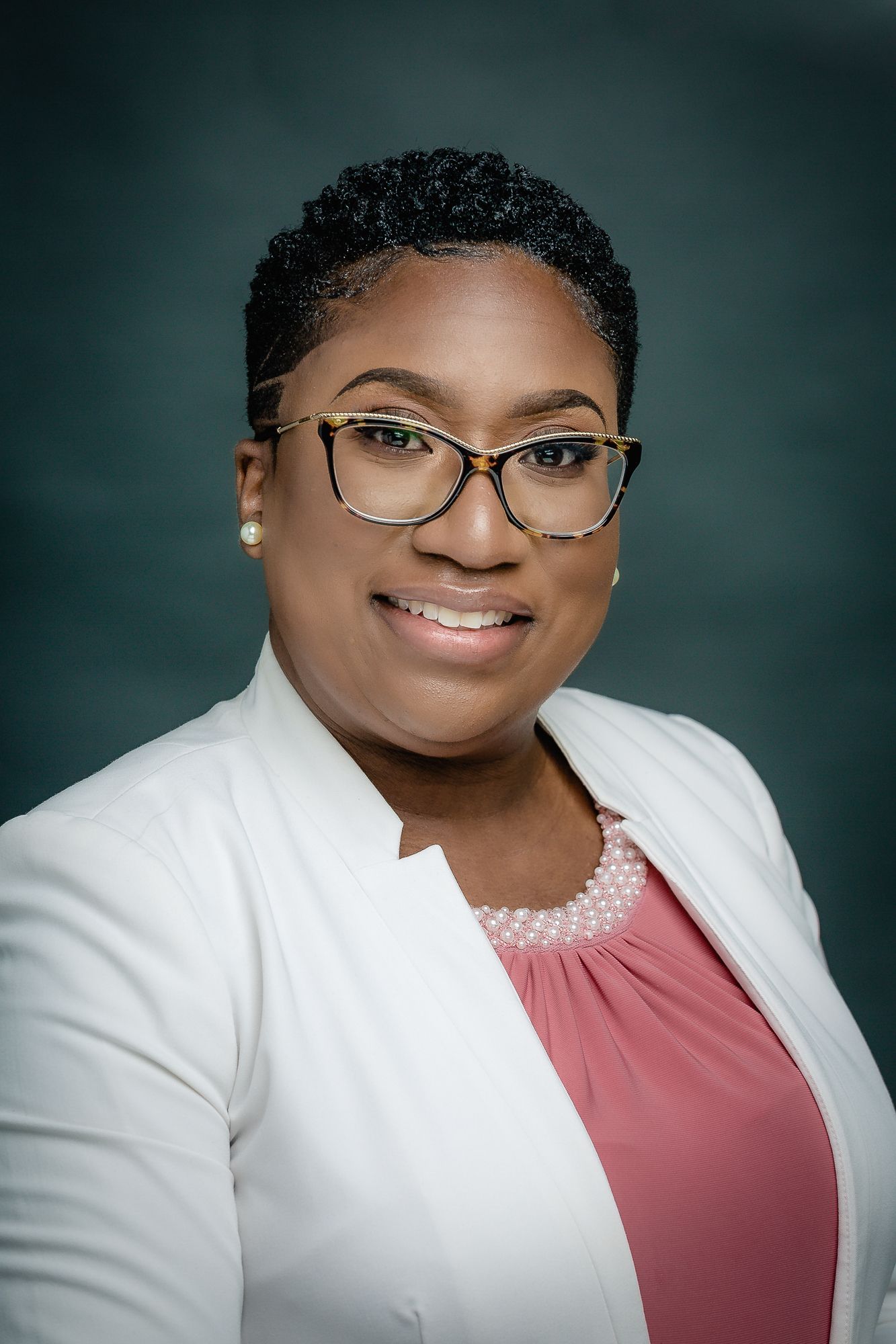 Professional headshot in white blazer and pink top with glasses against dark background.