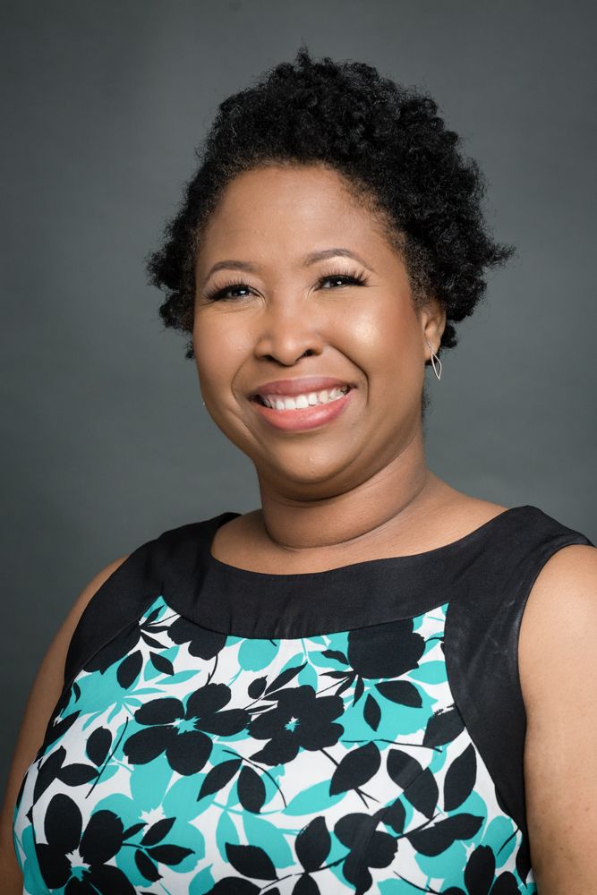 Professional headshot of someone wearing a sleeveless black and turquoise floral dress against a gray background.