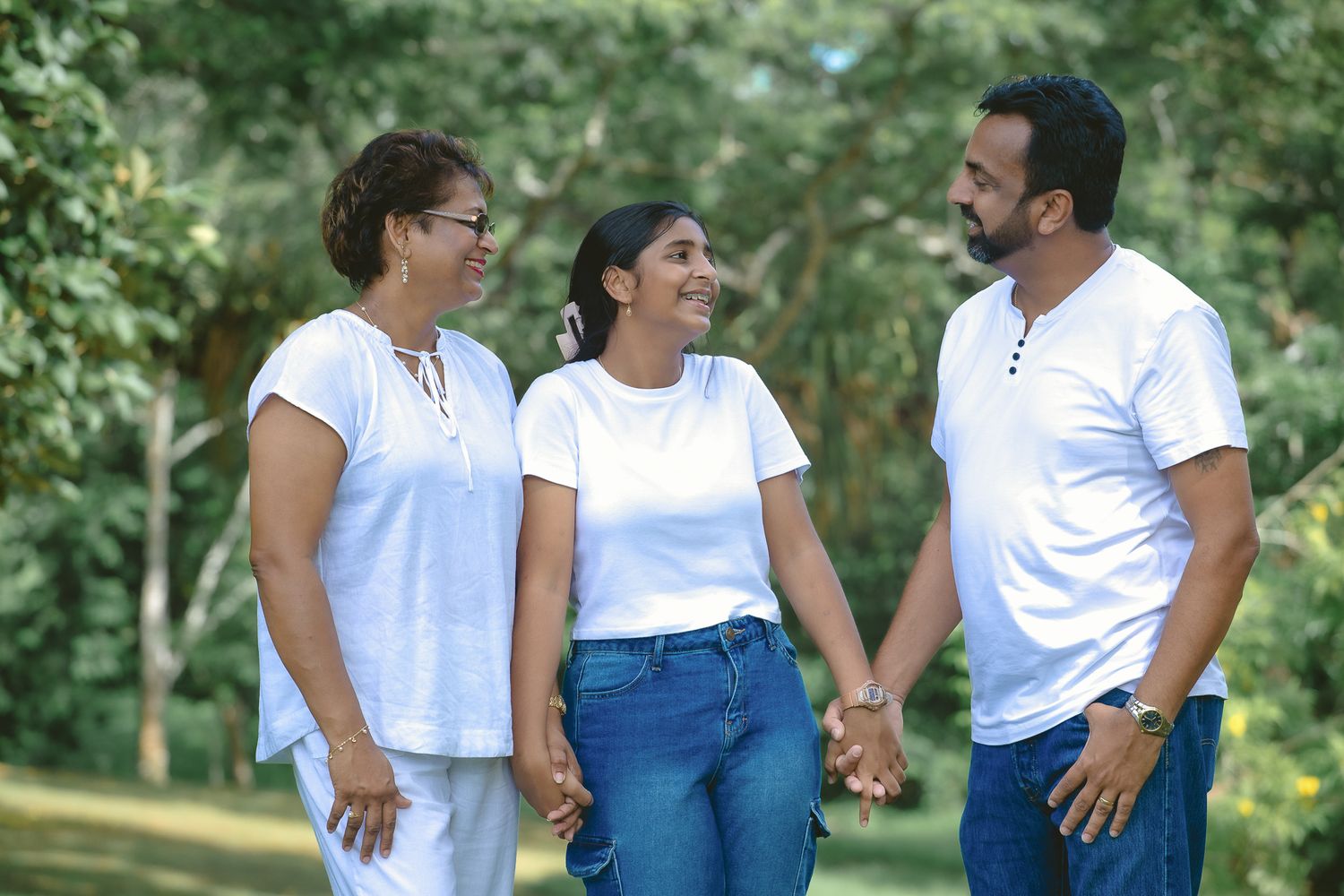 Three people wearing white tops and jeans walk together outdoors in a park on a sunny day.