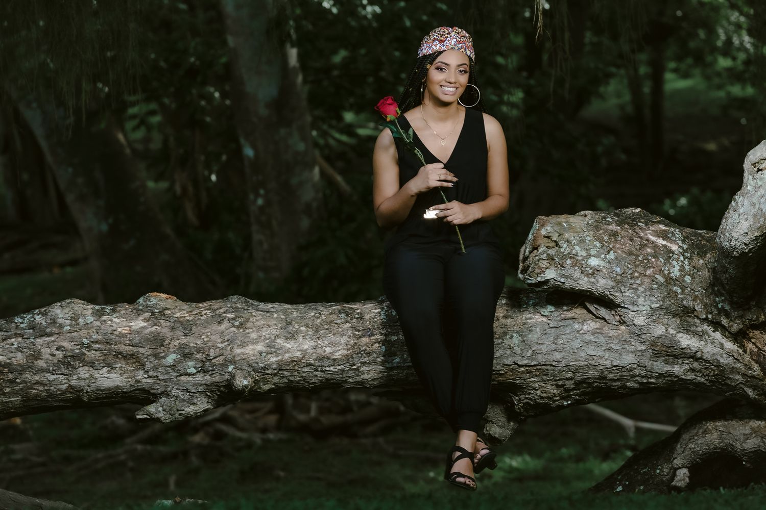 Woman in black dress poses with rose against rocky forest backdrop.