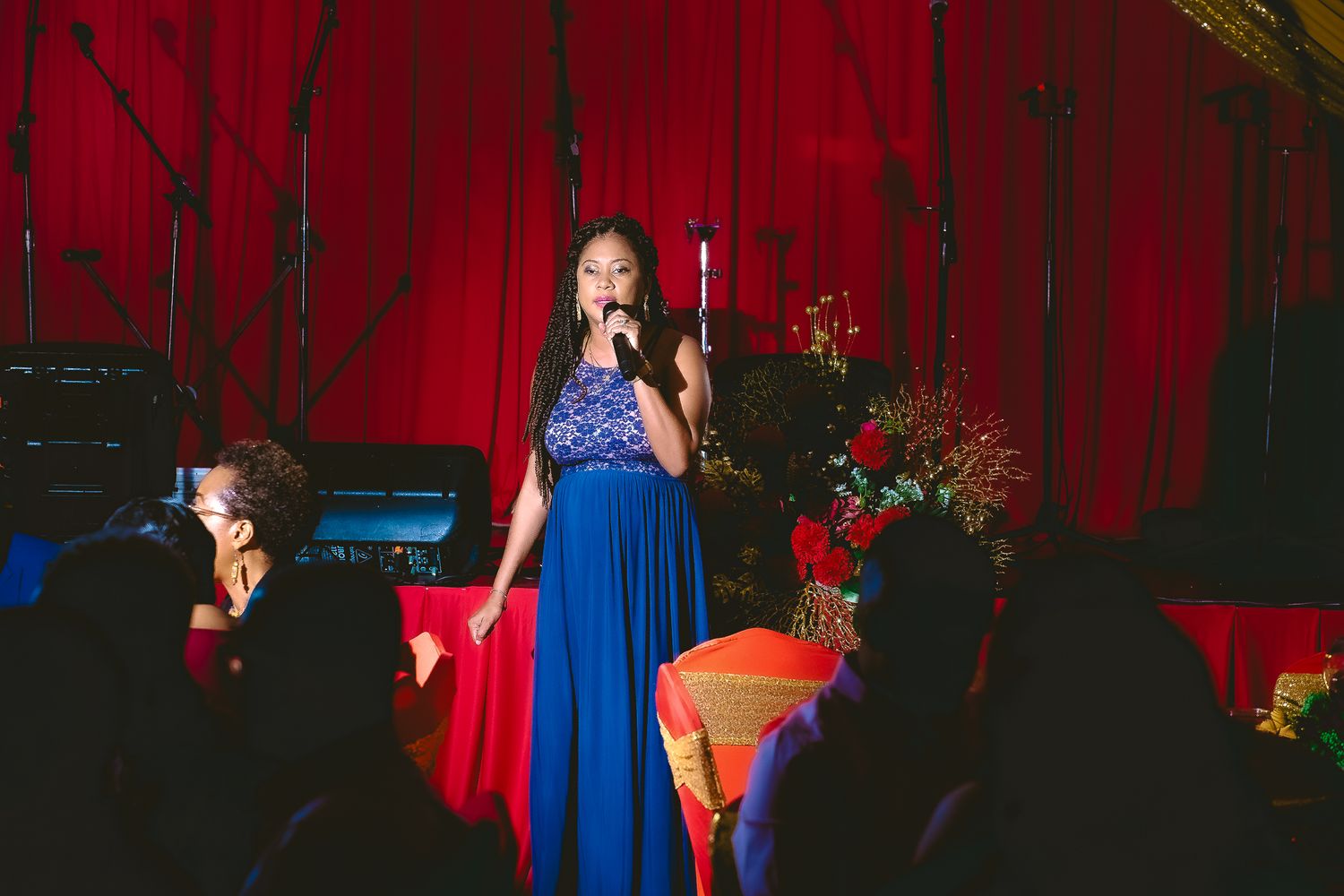 Performer in royal blue gown sings on stage with red curtain backdrop.