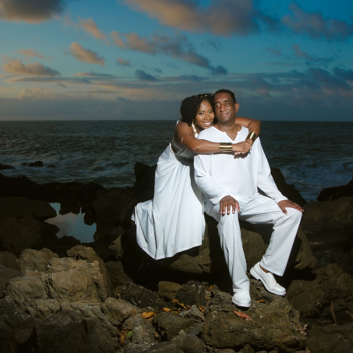 A romantic couple in white attire embraces while sitting on coastal rocks at sunset.
