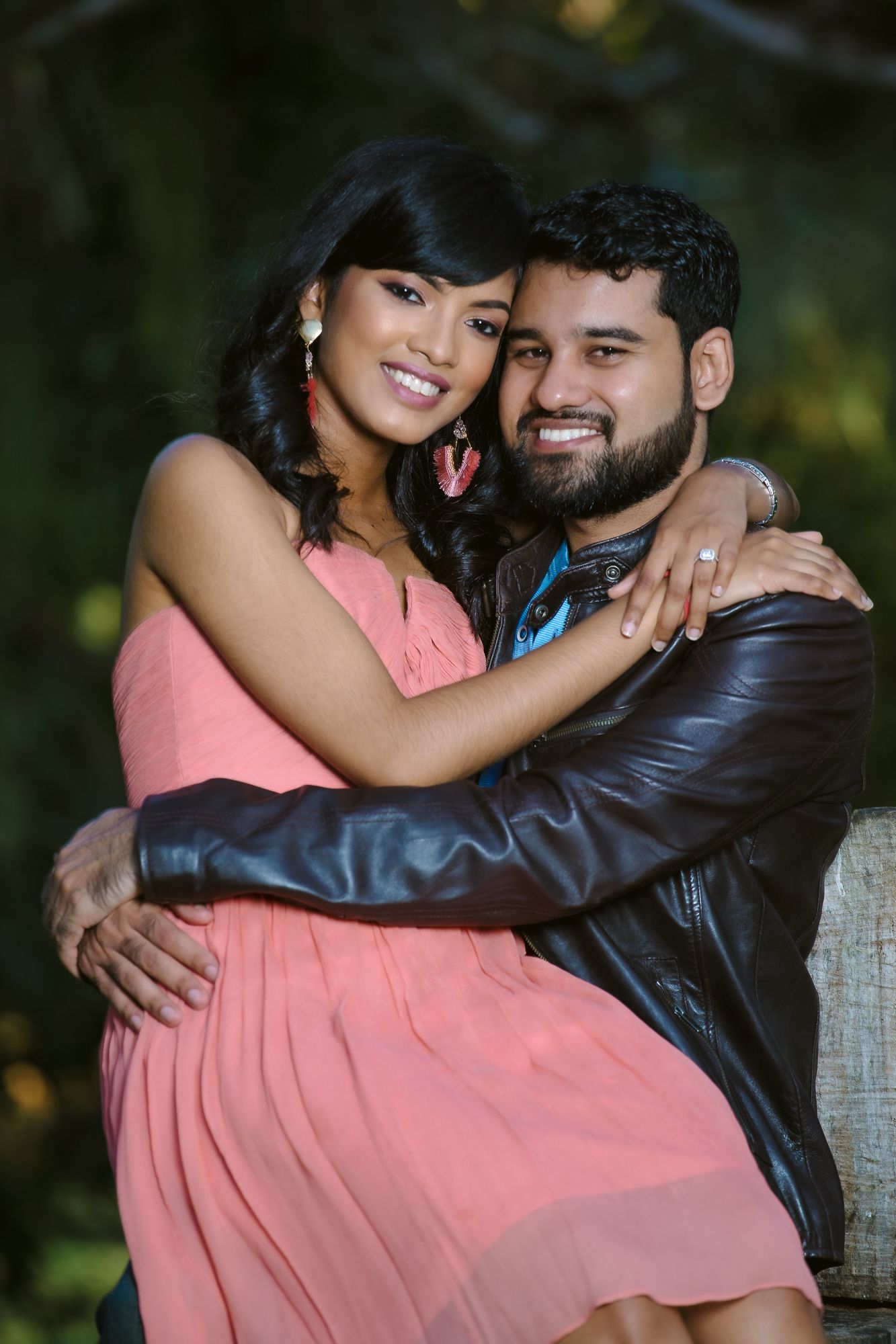 A couple embraces outdoors, with one wearing a pink dress and the other in a leather jacket against a natural background.