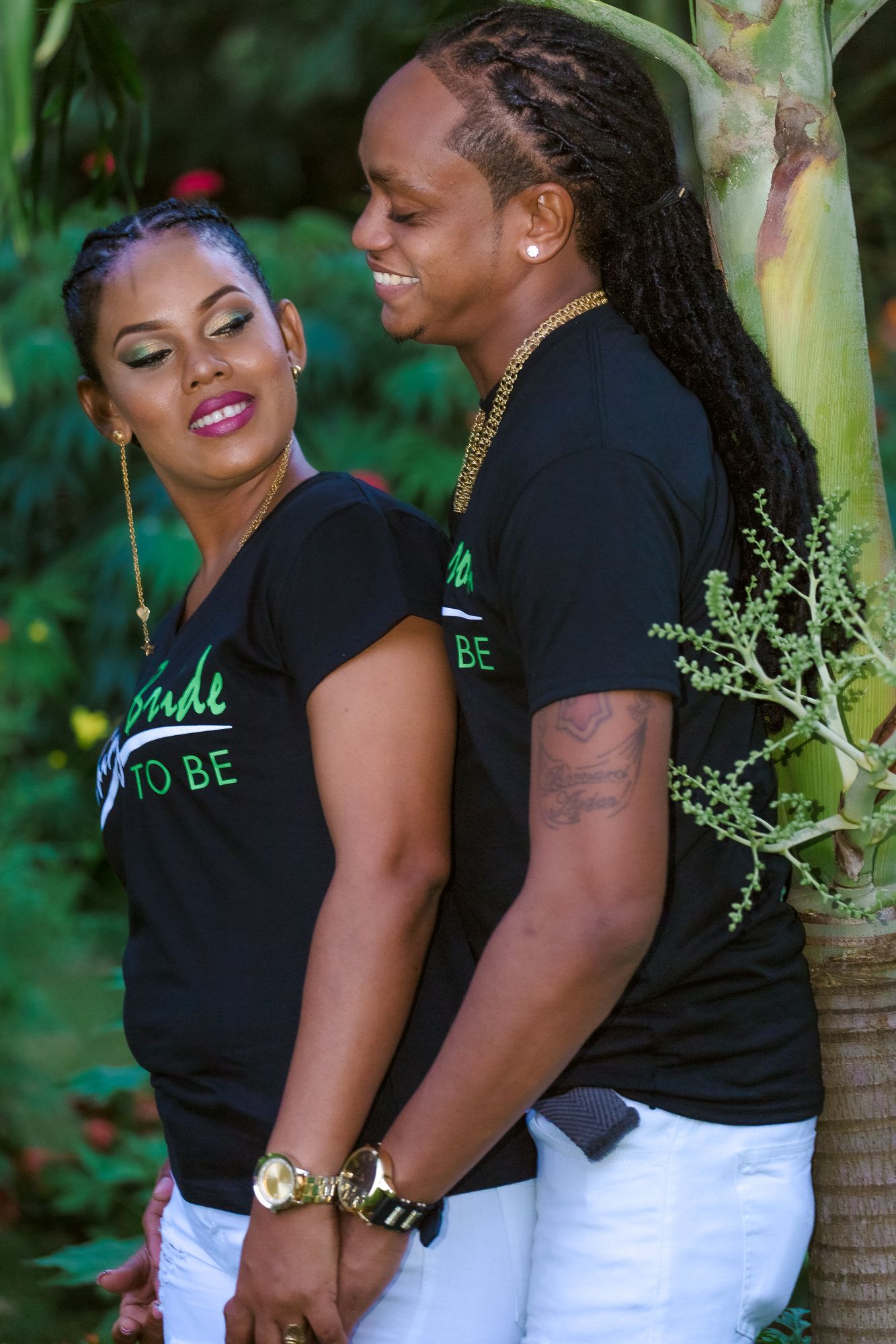 A couple in matching black t-shirts pose together against a tropical garden background.
