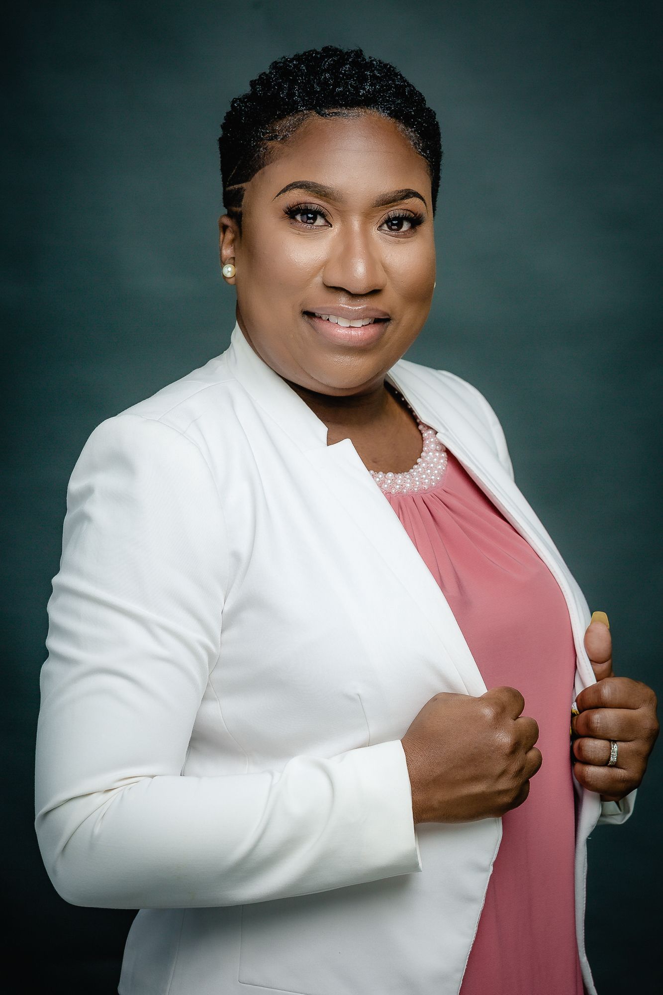 Studio portrait in white blazer and pink blouse against dark green background.