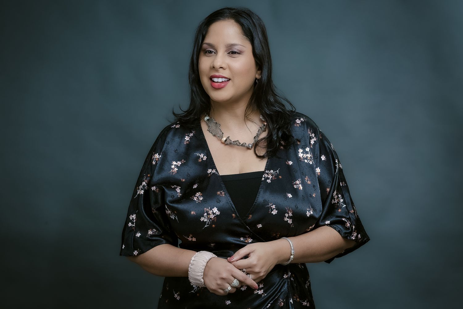 Studio portrait of a person in a floral dress with statement necklace against teal background.