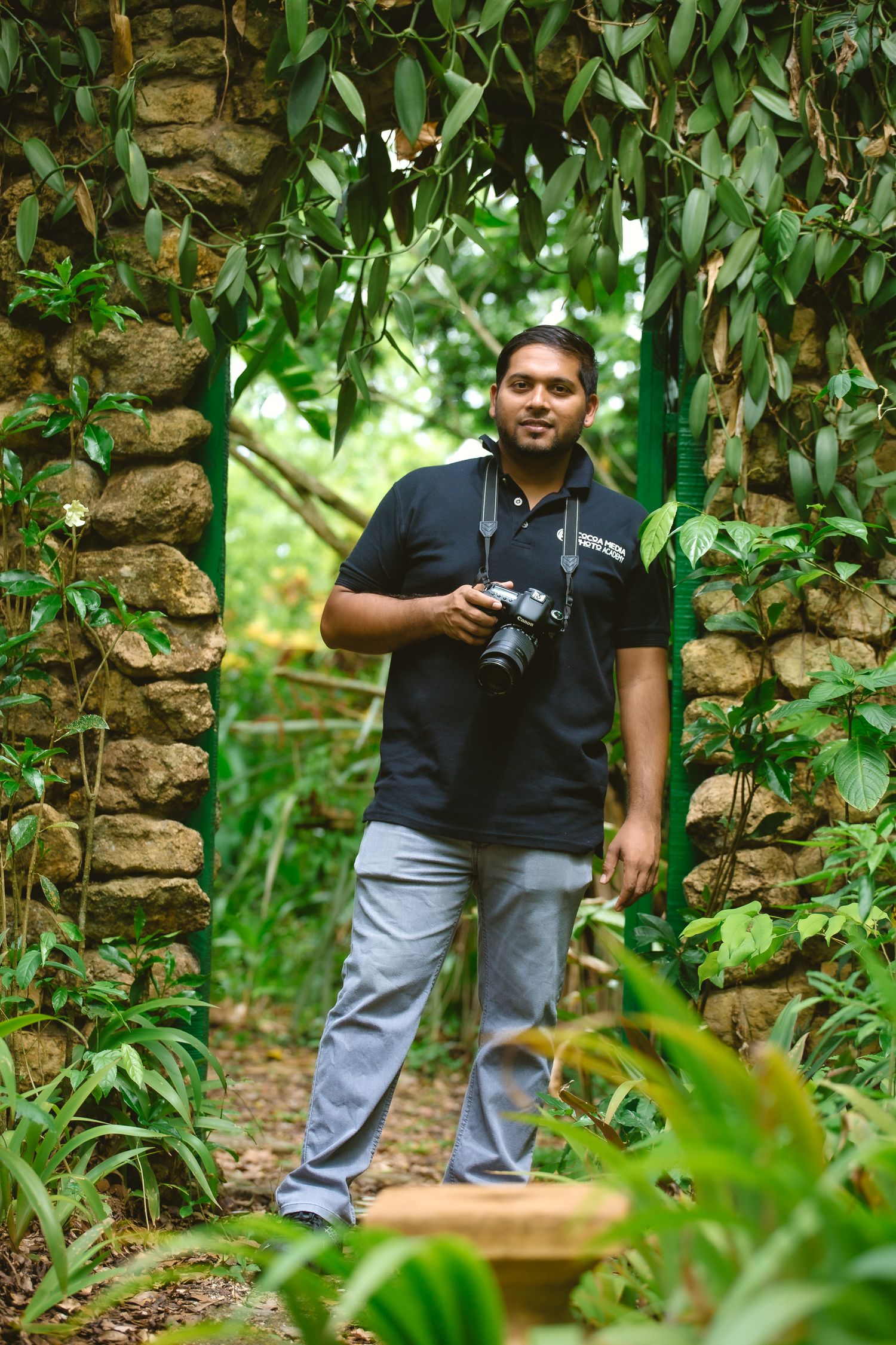 A photographer in a black polo shirt and gray pants stands among lush greenery and stone walls holding a camera.