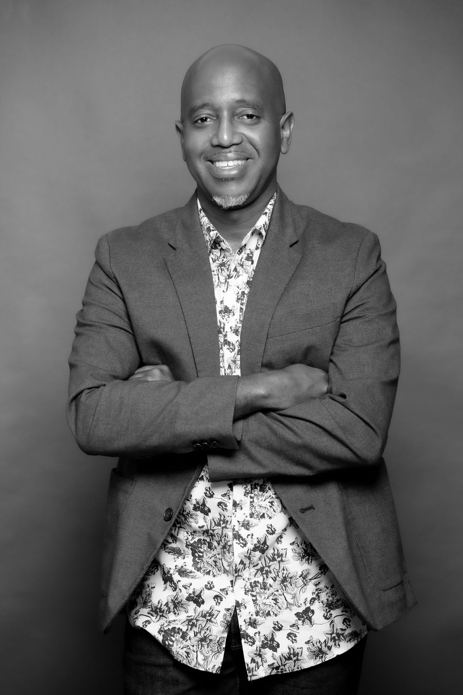 Professional headshot of a person in a blazer and floral shirt against a gray backdrop.