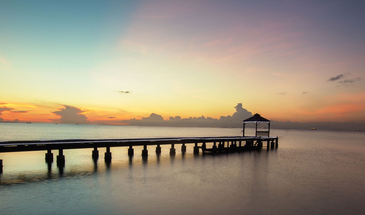 Long wooden pier stretches into calm waters during a peaceful sunset with a gazebo at the end.