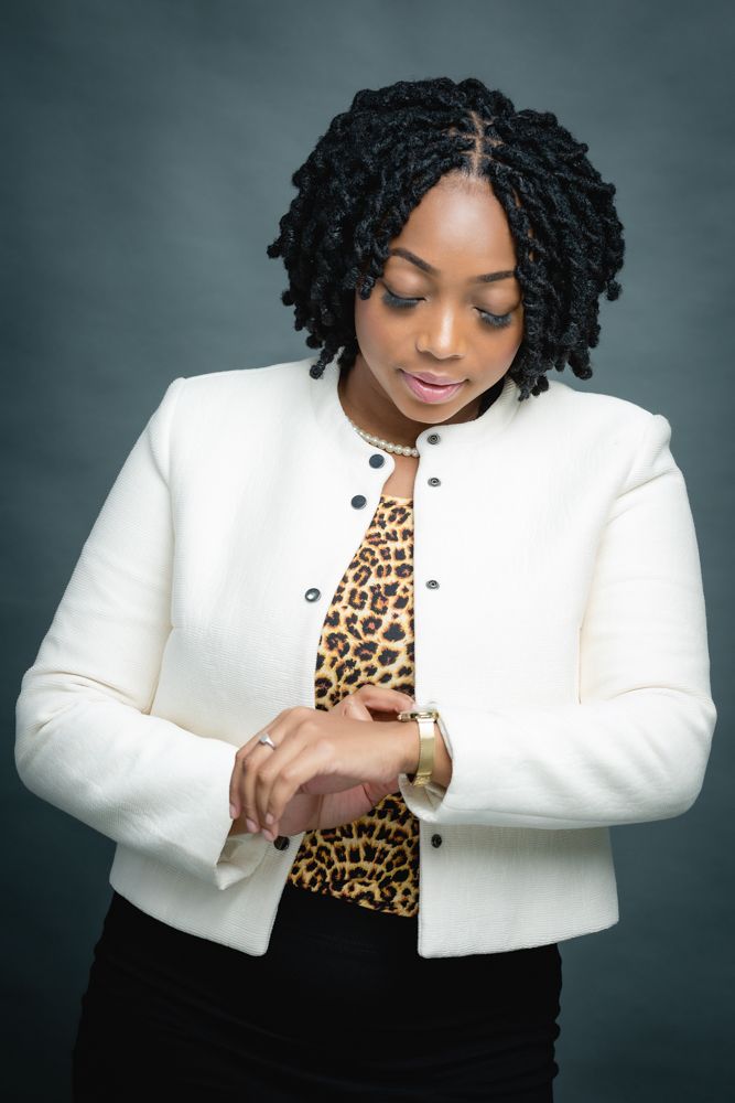 Professional portrait showing a person in a white blazer and leopard print top checking their watch against a dark background.