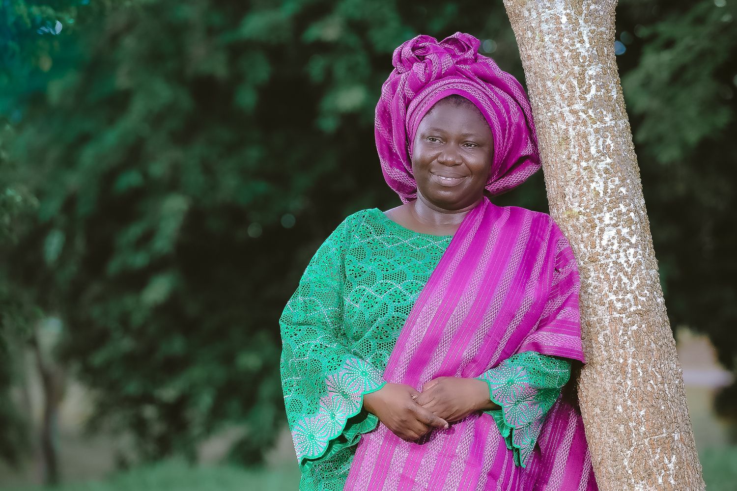 Woman in green lace and pink traditional African dress poses by tree.