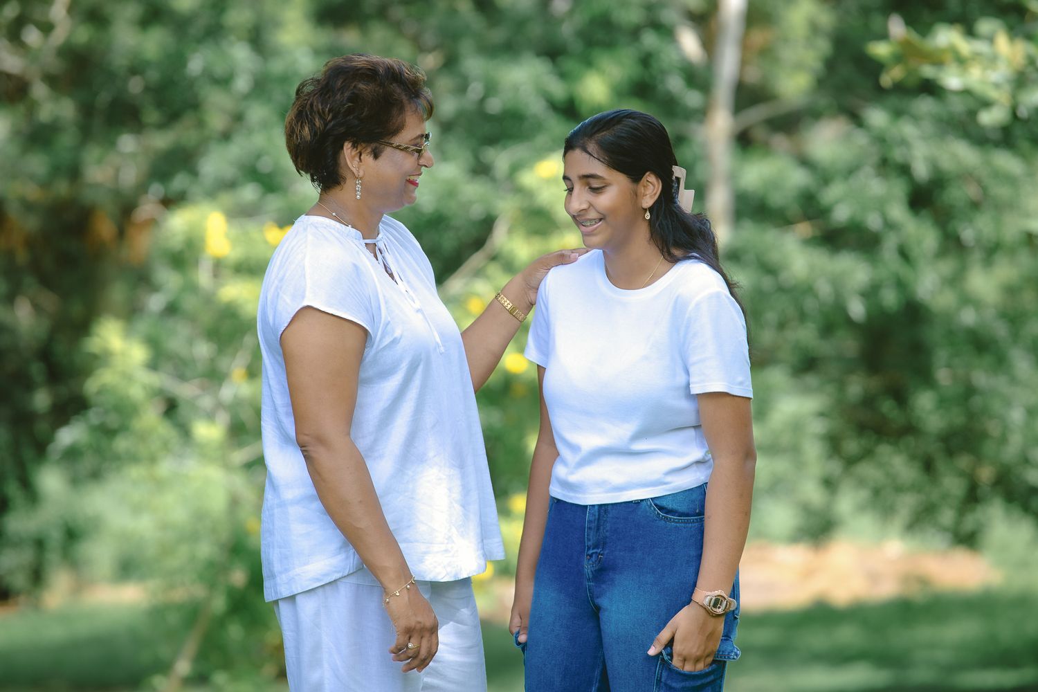 Two people in white t-shirts and casual attire share a conversation in a leafy outdoor setting.