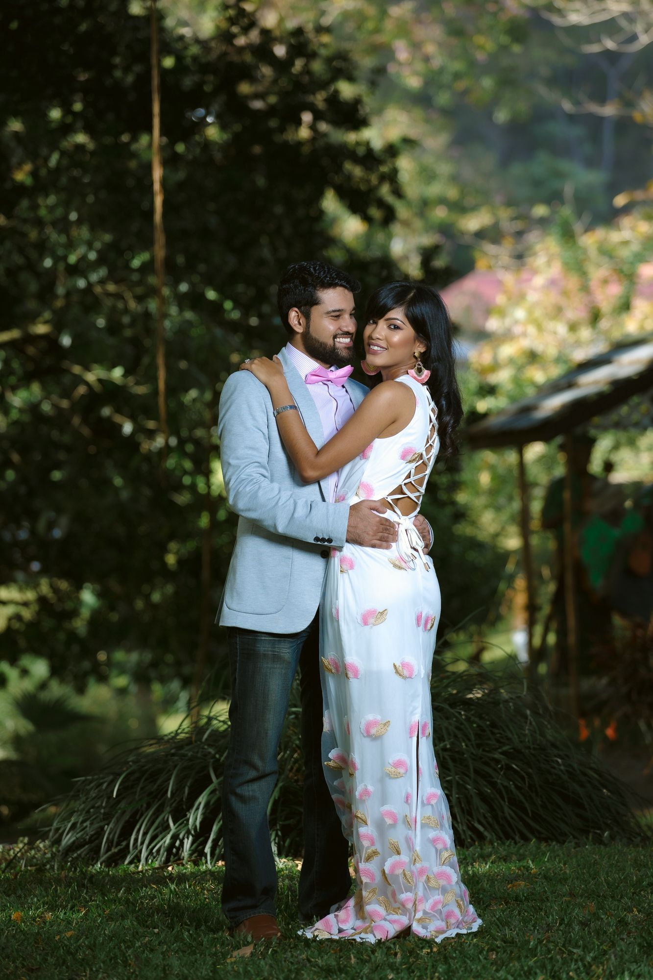 A romantic moment captured outdoors of a couple in formal attire embracing on a garden lawn with lush greenery behind them.