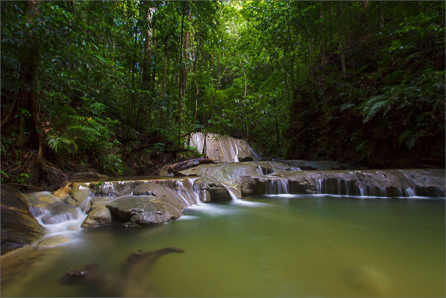 Serene waterfall cascades over rocks into green pool in lush tropical forest.
