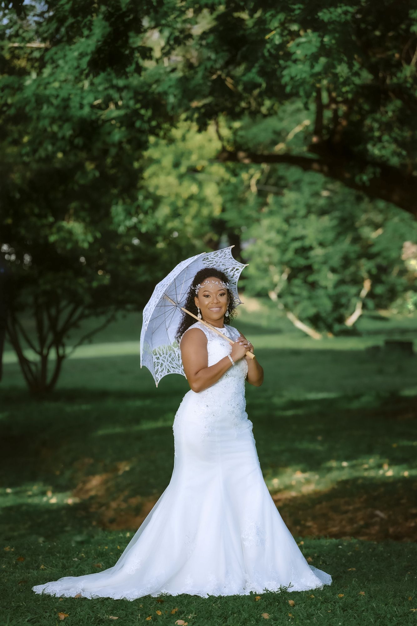 A bride in a fitted mermaid wedding dress poses with a white lace parasol in a lush garden setting.