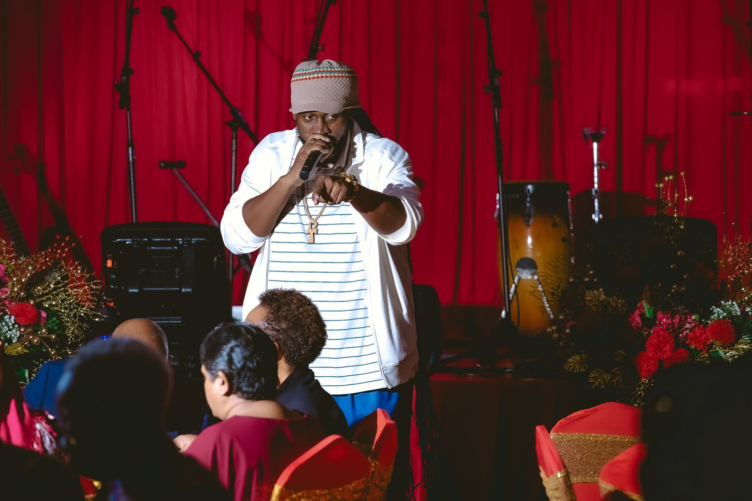 Performer in white shirt singing into microphone on stage with red curtain backdrop.