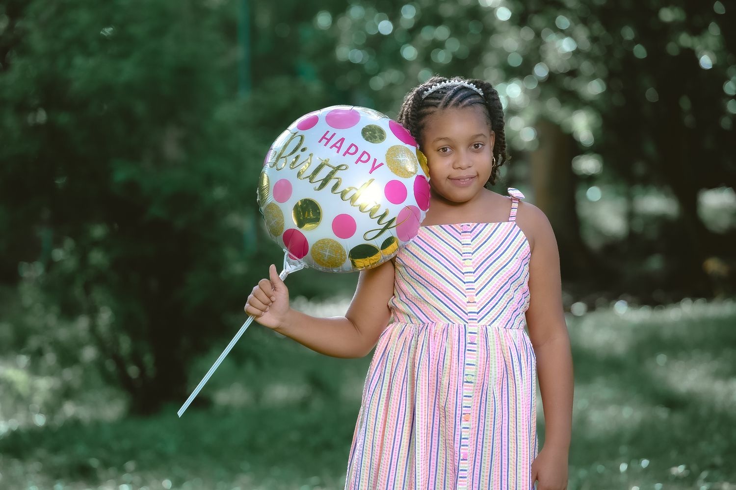 Child in striped dress holds colorful polka dot birthday balloon in outdoor garden setting.