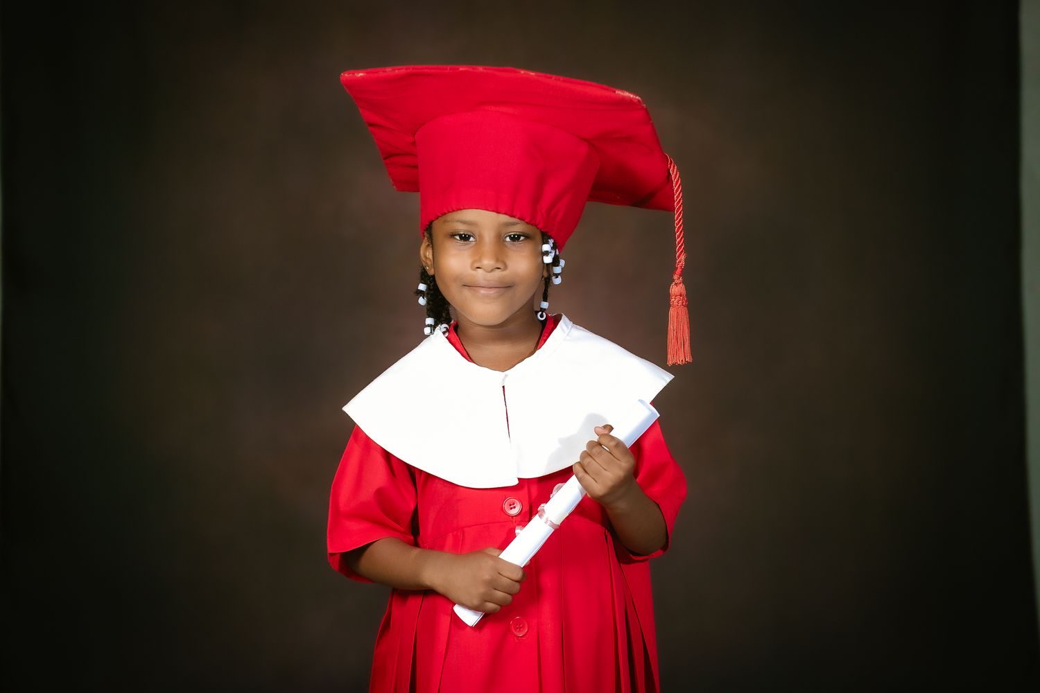 Child in red graduation cap and gown poses with diploma during preschool graduation photo session.