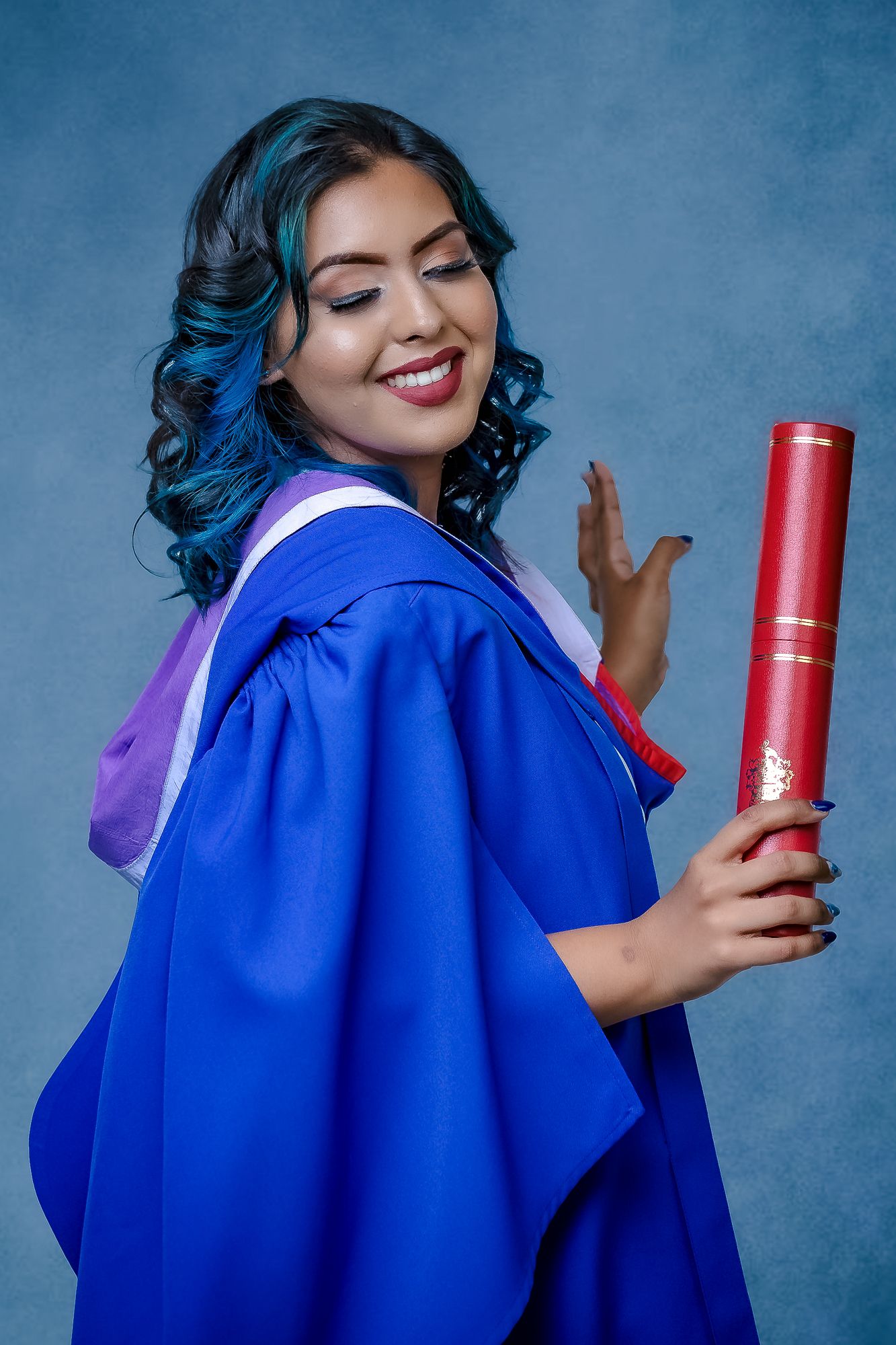 Graduate in blue academic regalia with blue hair holding red diploma case.