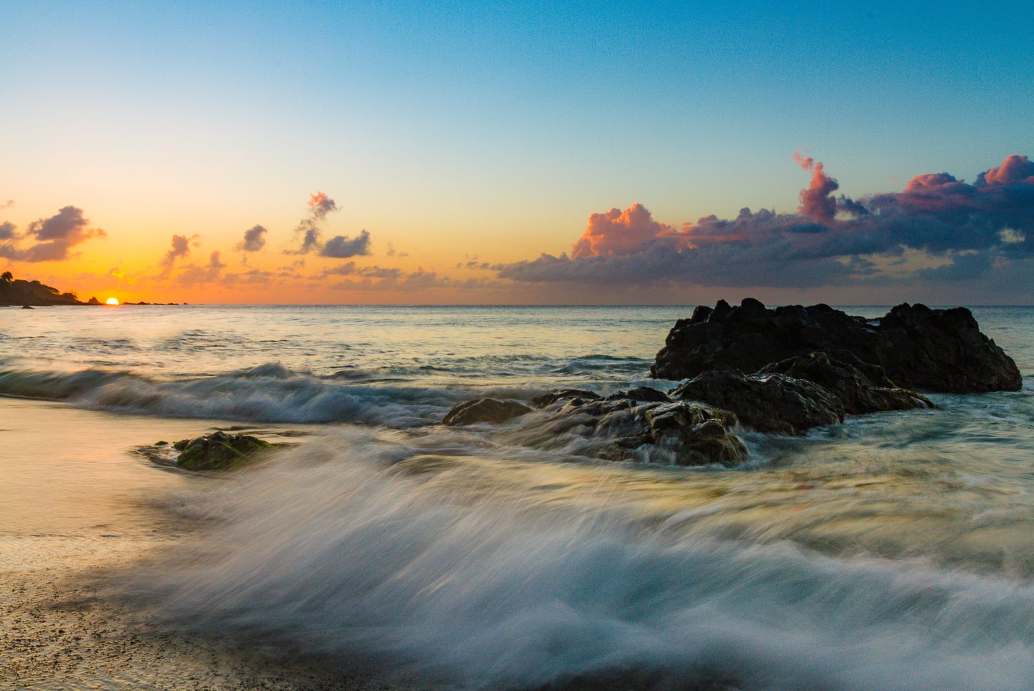 Dramatic coastal sunset with waves rushing over rocks at beach during golden hour.