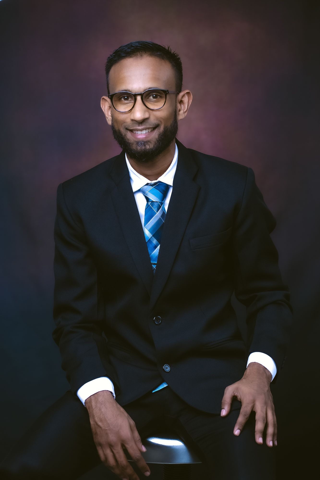 Professional portrait of person in suit and striped tie seated against dark background.