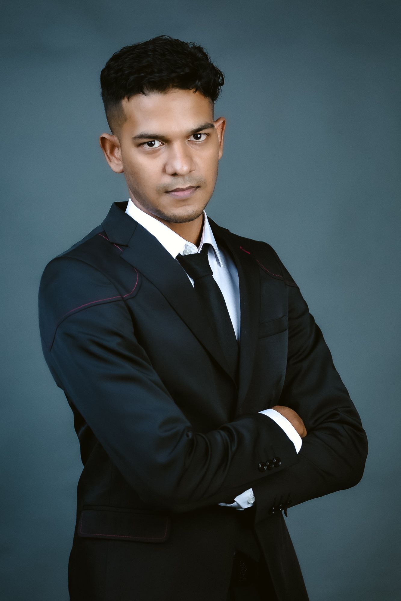 Professional portrait of a person in black suit with crossed arms against blue background.