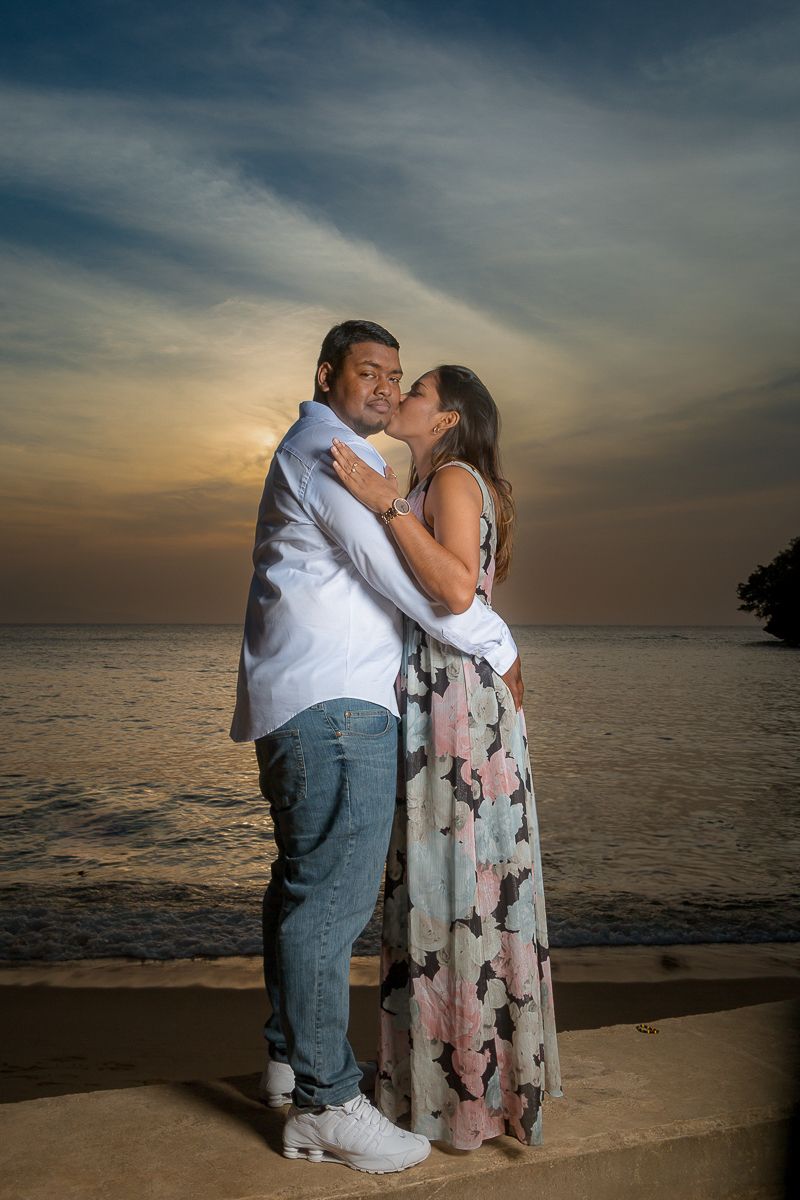 A couple embraces in a romantic sunset moment by the ocean, silhouetted against a dramatic sky with wispy clouds.