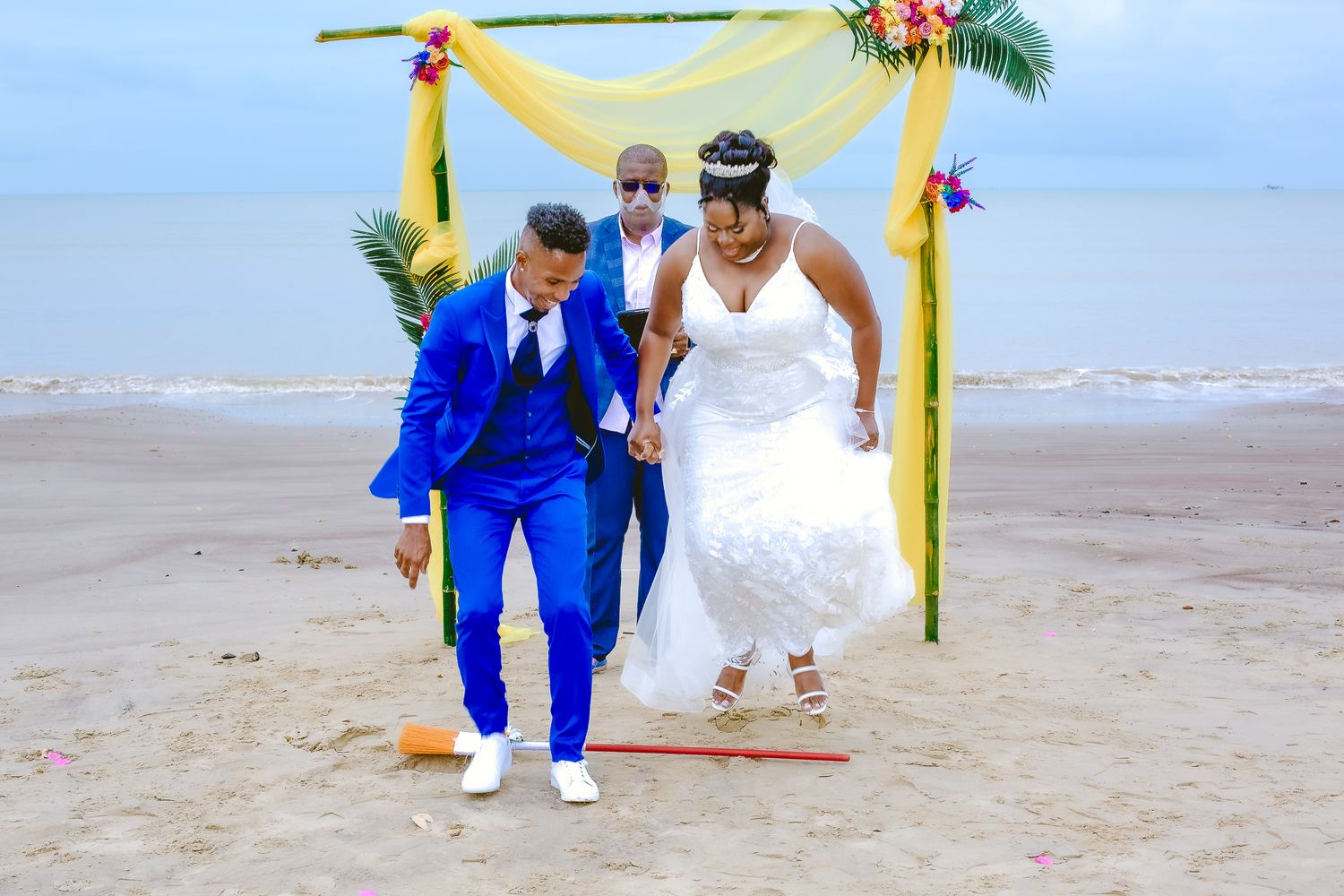 Newlyweds jumping over a broom on beach under yellow arch with flowers.