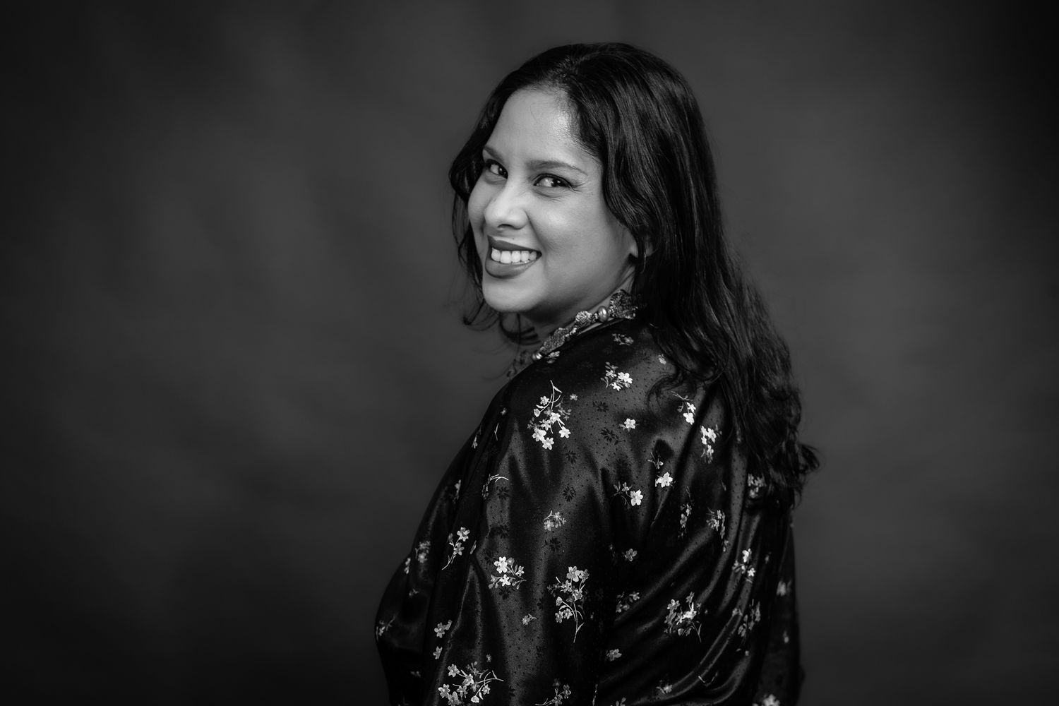 Black and white portrait of a person in floral dress smiling over their shoulder.