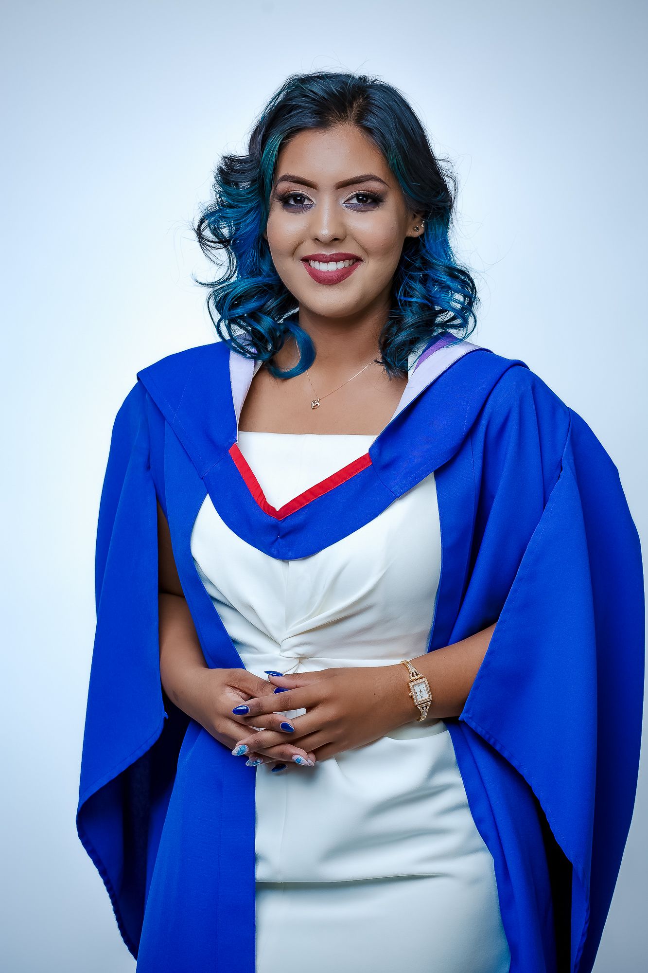 Graduate in blue academic dress and white outfit poses confidently against light background.