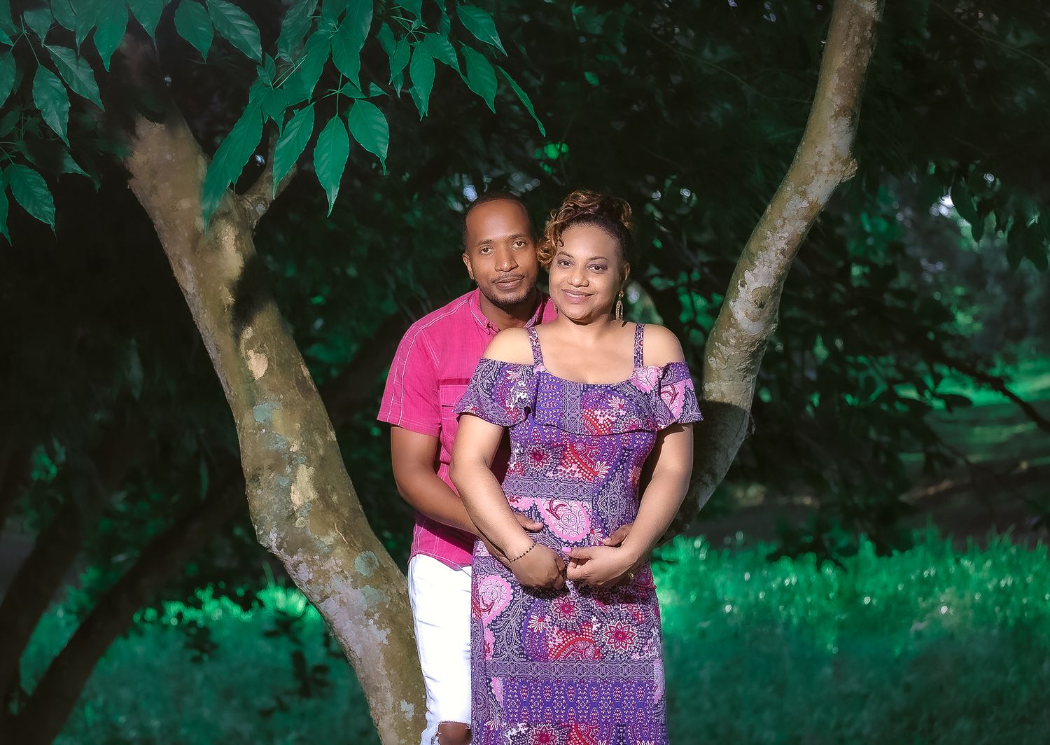 A couple poses together in purple and pink outfits against a backdrop of green trees.