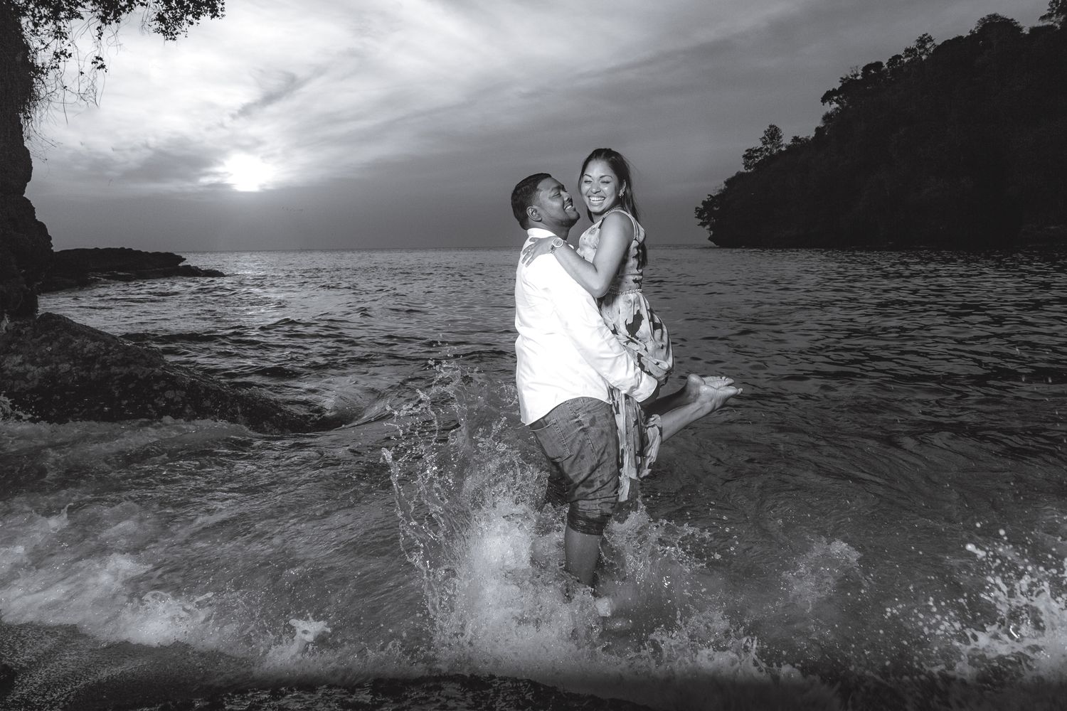 A romantic black and white photograph of a couple embracing in ocean waves at sunset along a rocky coastline. Captured at Macqueripe