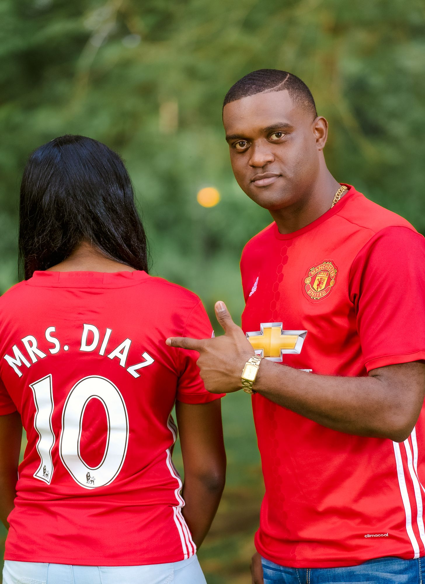 A couple wearing matching red Manchester United jerseys poses together in an outdoor setting with green foliage.