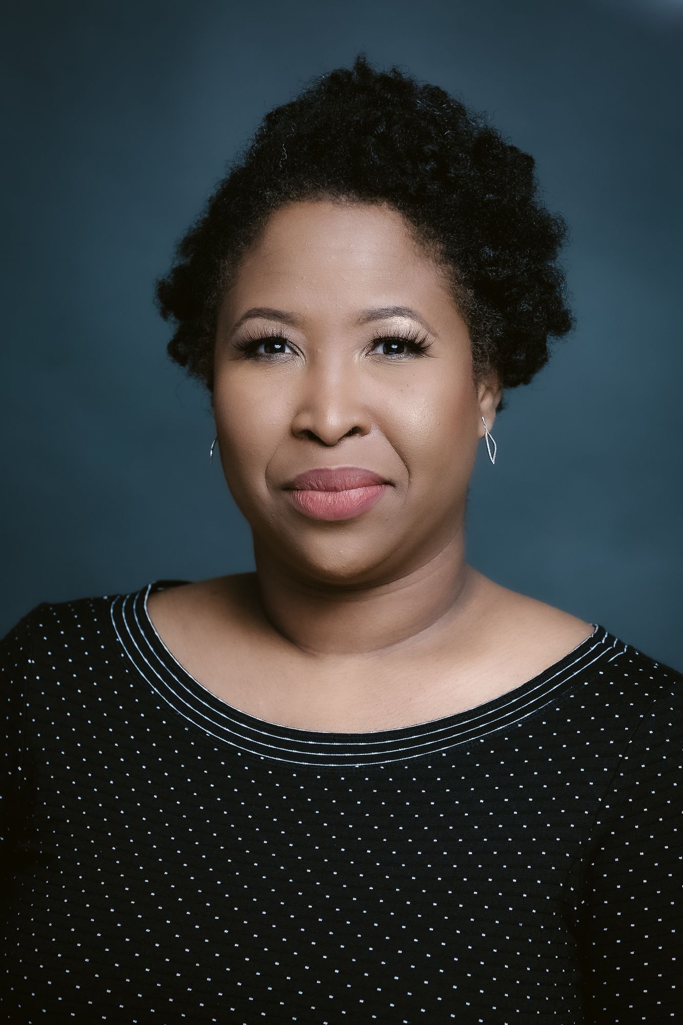 Professional studio portrait showing confident pose in black polka dot top against dark background.