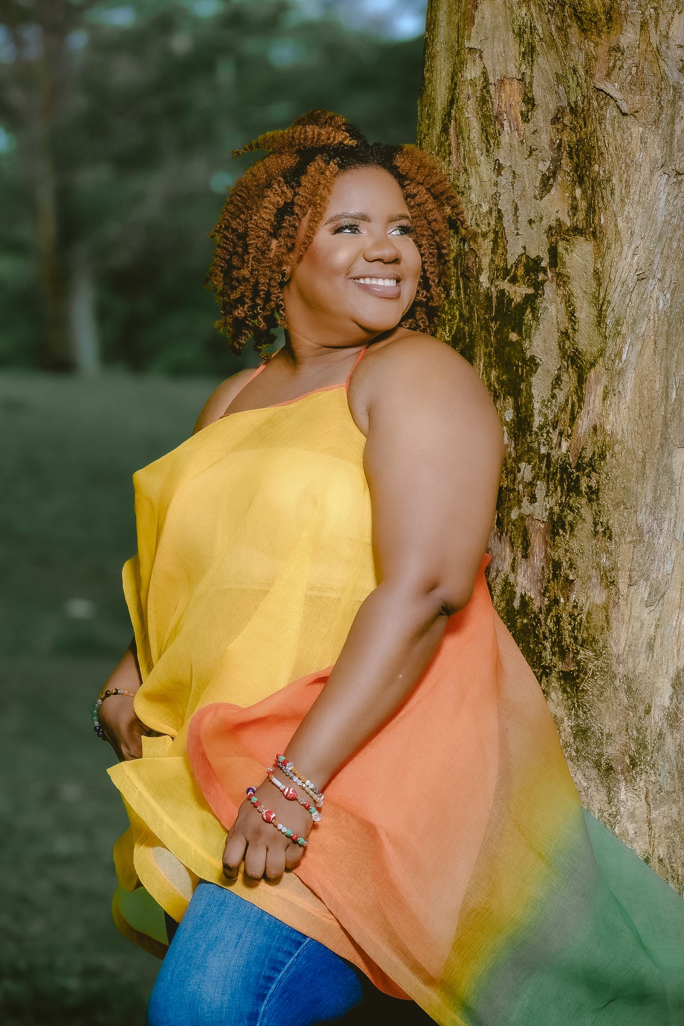 Woman in yellow and orange ombre top leaning against tree trunk.