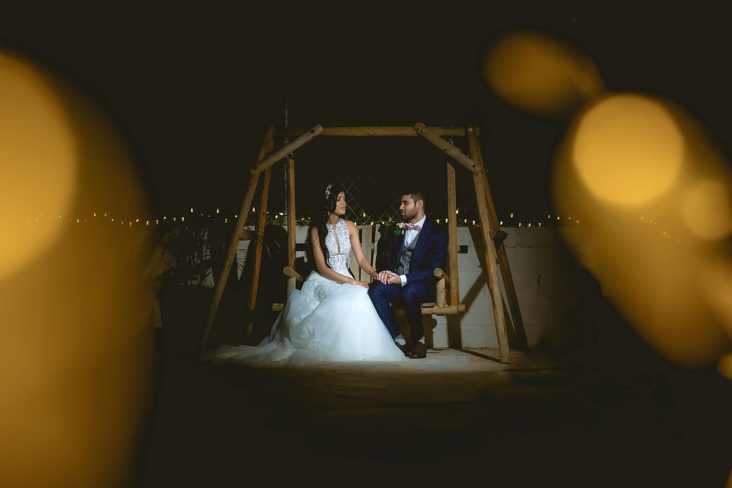 A wedding couple sits on a swing at night with bokeh lights in the background.