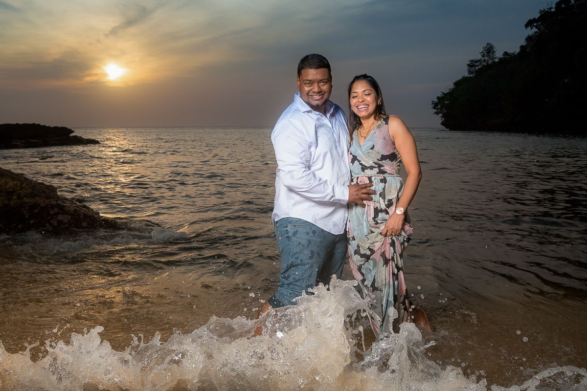A couple stands in splashing waves on a beach at sunset with dramatic sky and rocky coastline in the background.