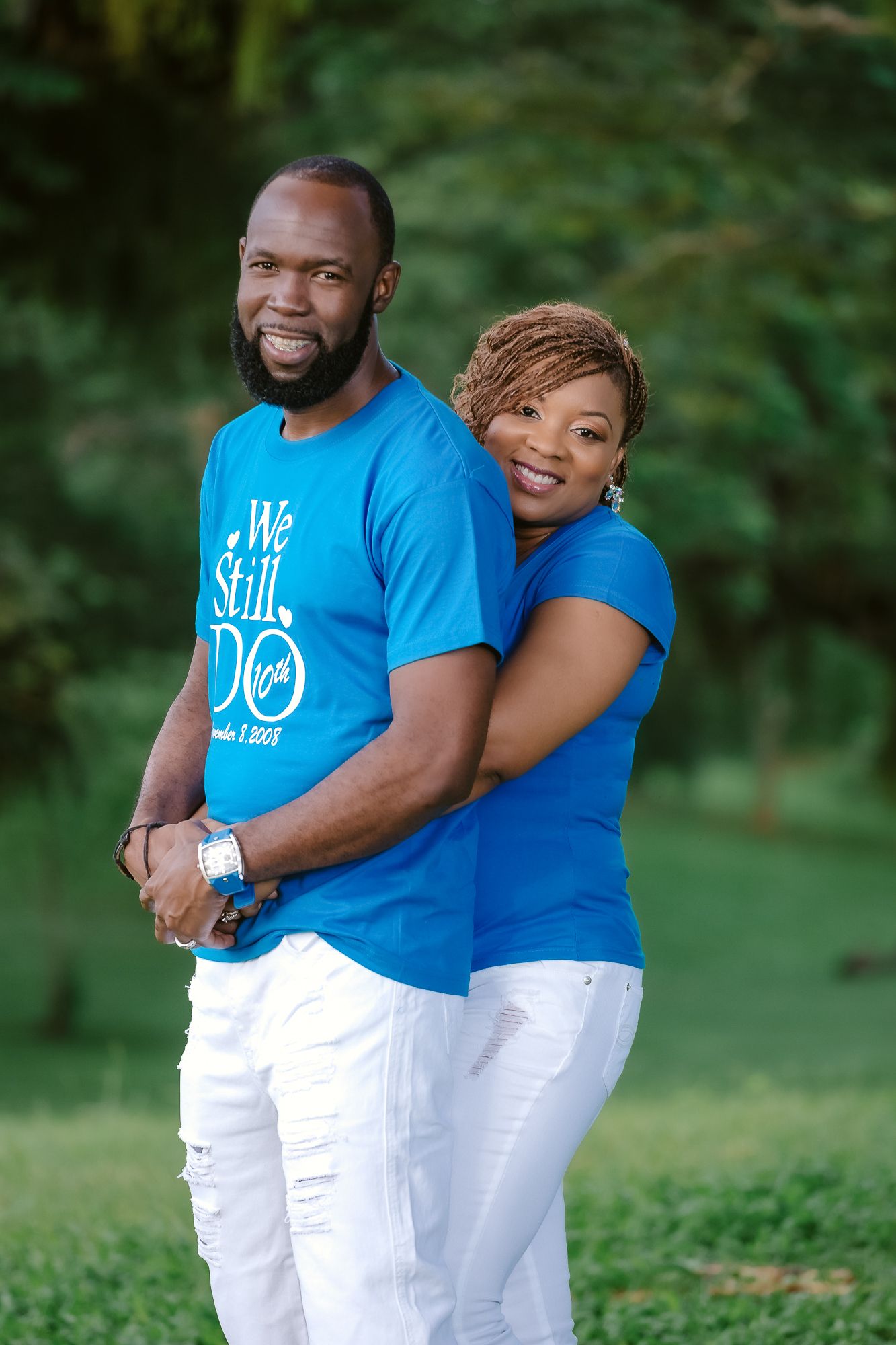 A couple in matching blue shirts and white pants pose together in a park setting.