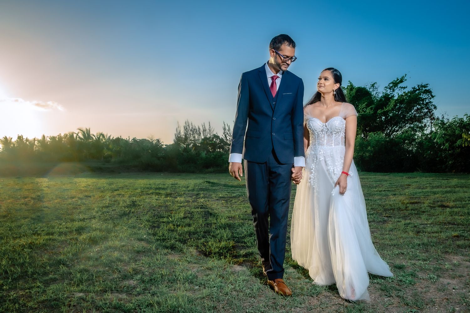 Newlywed couple walking together at sunset in an open field wearing elegant wedding attire.