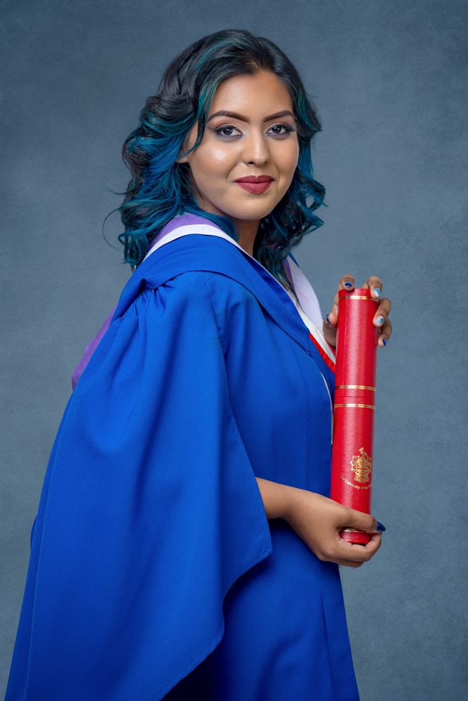A person in a royal blue graduation gown poses with a red cylindrical container against a gray backdrop.