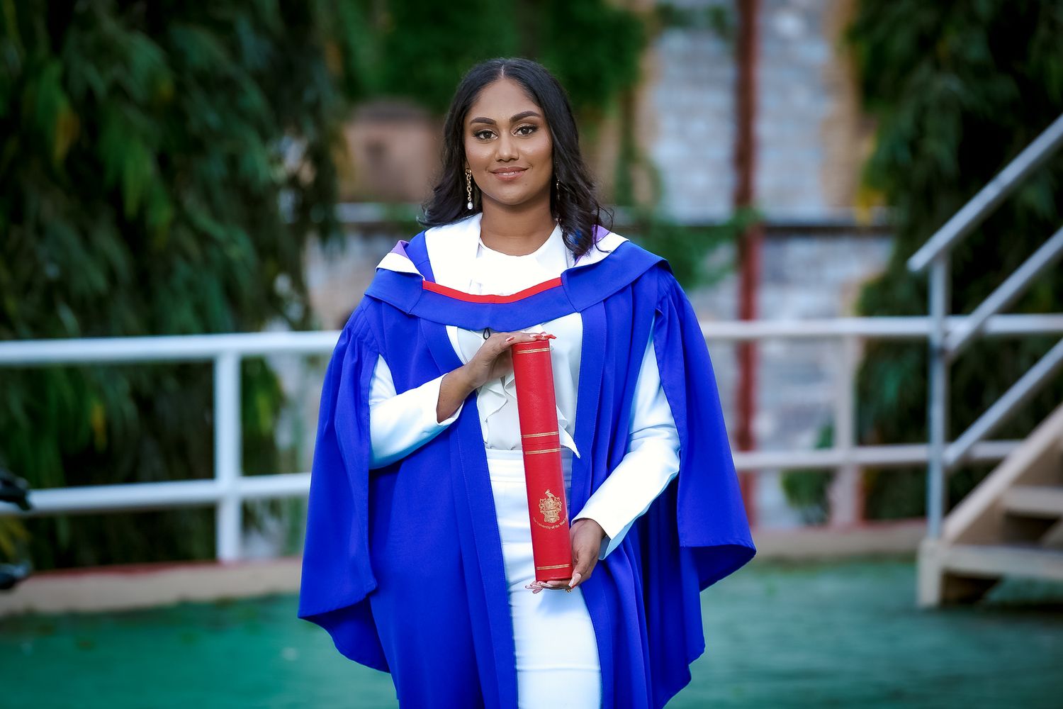 Graduate in blue academic regalia poses outdoors on campus with red and white accents.